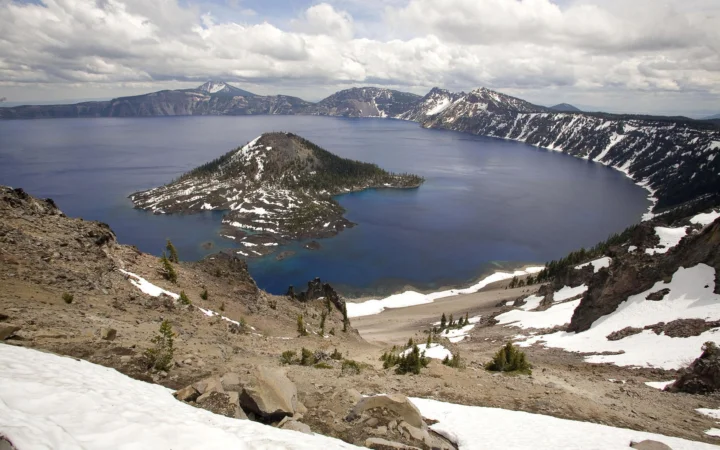 一片深蓝色的火山湖坐落在积雪山峰之间，湖心有一座绿树覆盖的小岛，天空布满云层。