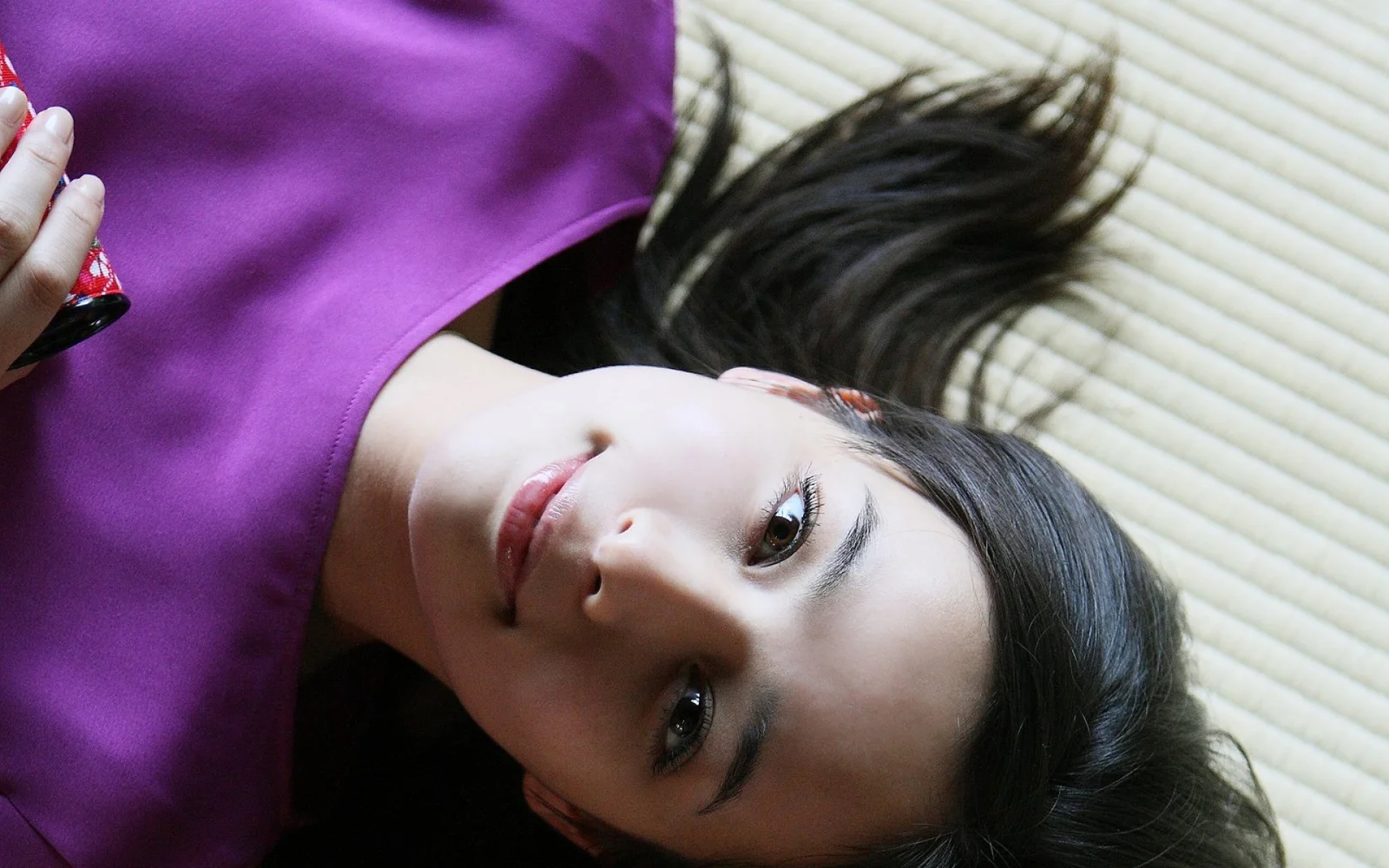 一名穿紫色上衣、留着深色长发的成年女性侧躺在百叶窗前。