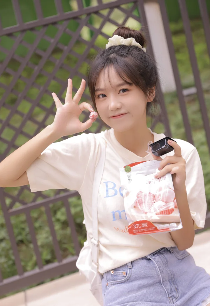 お団子ヘアの女性が木製の格子フェンスの前で微笑み、スナックの袋と黒い小物を手にしています。