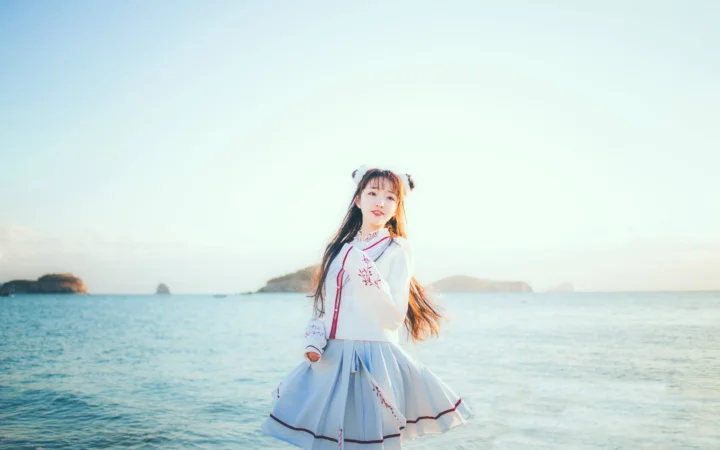 一名身穿浅色连衣裙的女子站在海边，远处可见海岛和开阔海面。