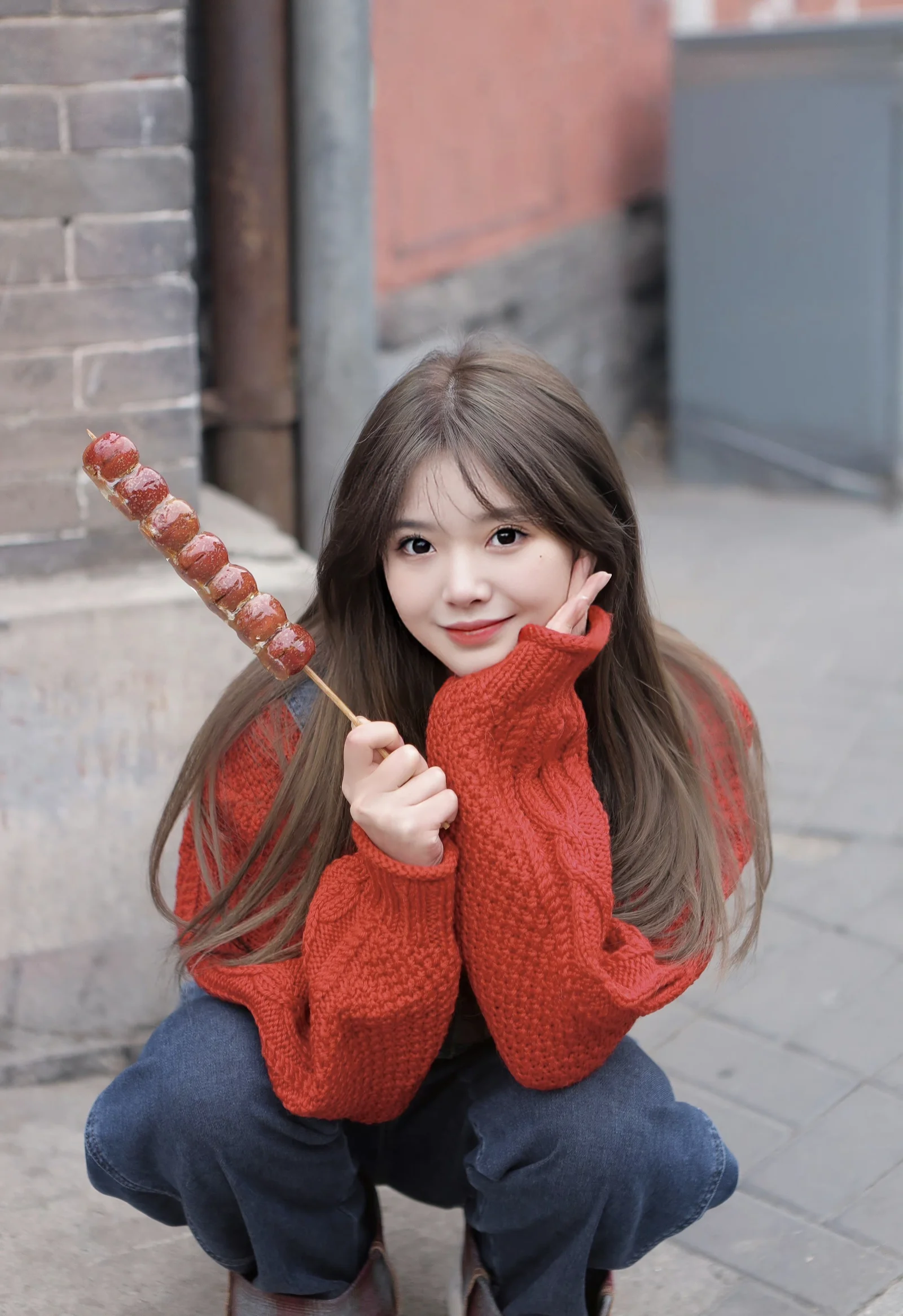 빨간 스웨터를 입은 긴 머리 여성이 달콤한 과일 꼬치를 들고 거리에서 카메라를 향해 미소 짓고 있다.