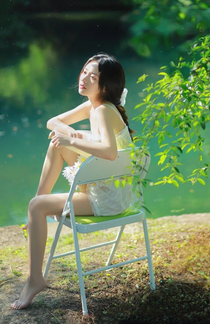 一名身穿白色连衣裙的女子坐在绿水和树木旁的折叠椅上。