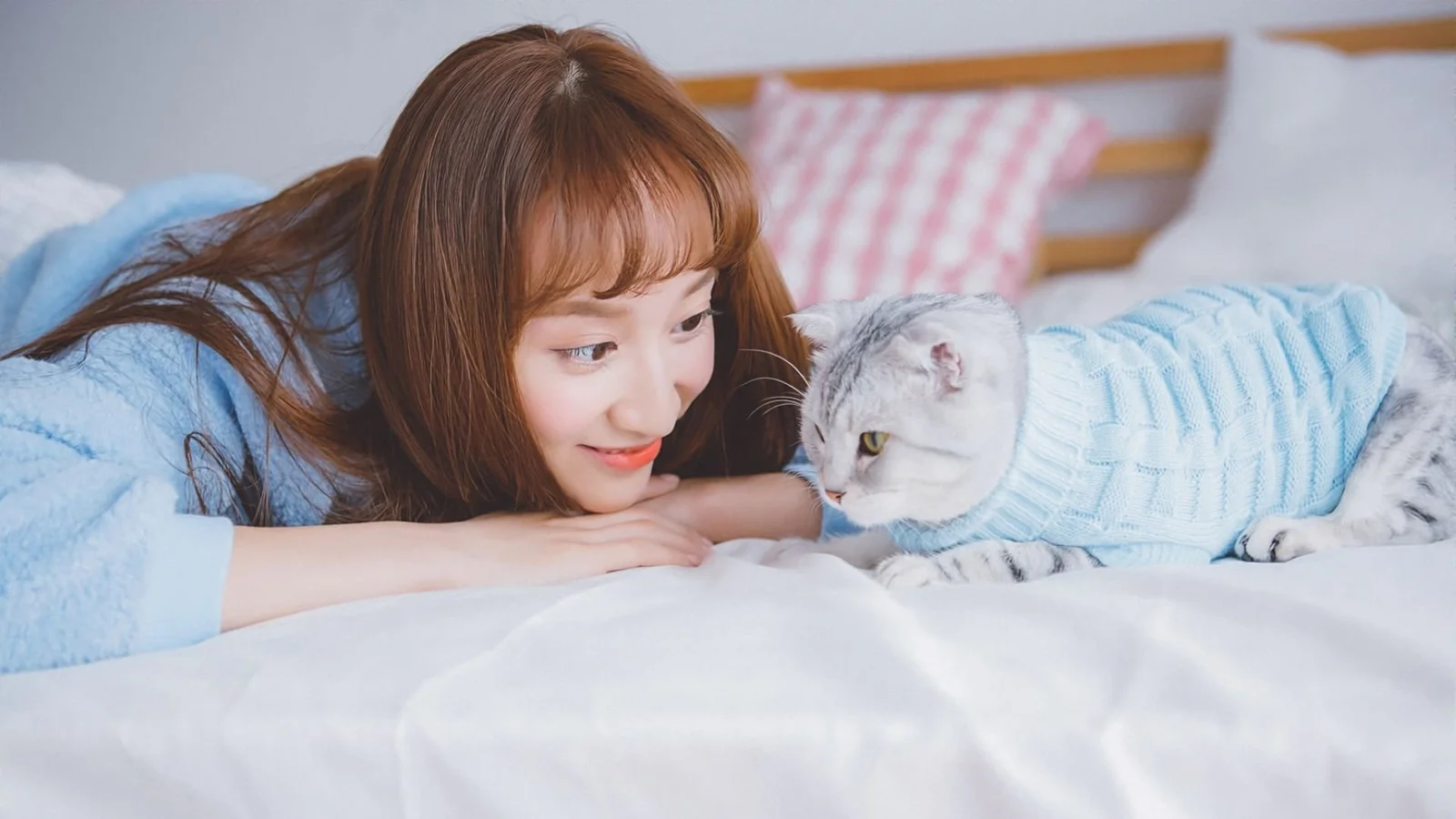 한 여성이 침대 위에서 연한 파란색 니트 옷을 입은 고양이와 서로 바라보고 있다.