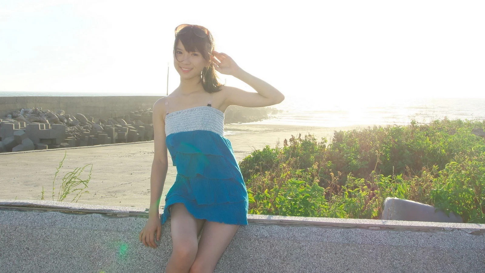 一名女子坐在海边的堤岸上，阳光照亮了海面和周围植被。