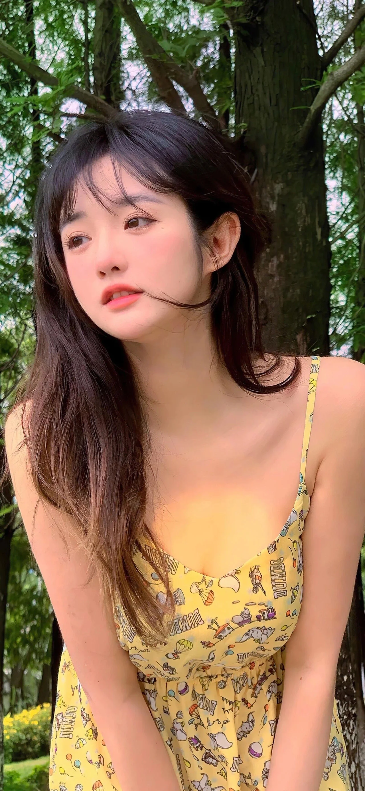一位长发女子身穿黄色图案连衣裙站在树木间。