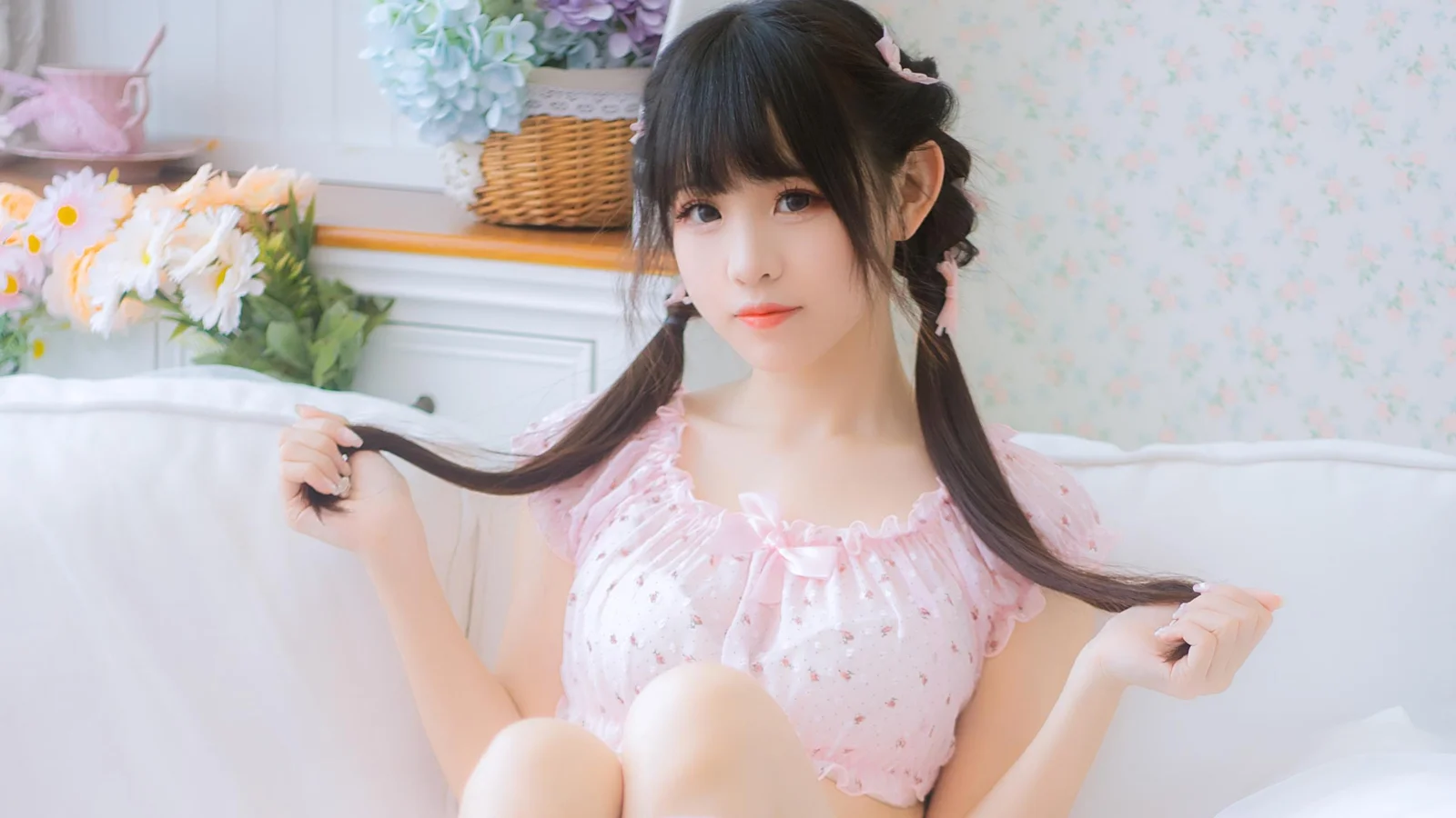 一名长发女性身穿粉色上衣坐在明亮的室内环境中，手持两侧发束面向镜头。