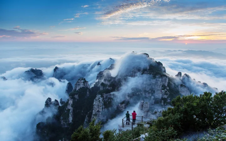 云雾缭绕的群山延伸在日出天空下，前景观景台上可见两名远处行人。