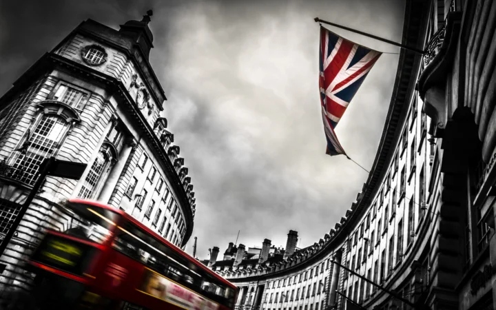 黑白建筑环绕的英国城市街道上，一辆红色巴士驶过，空中悬挂着英国国旗。