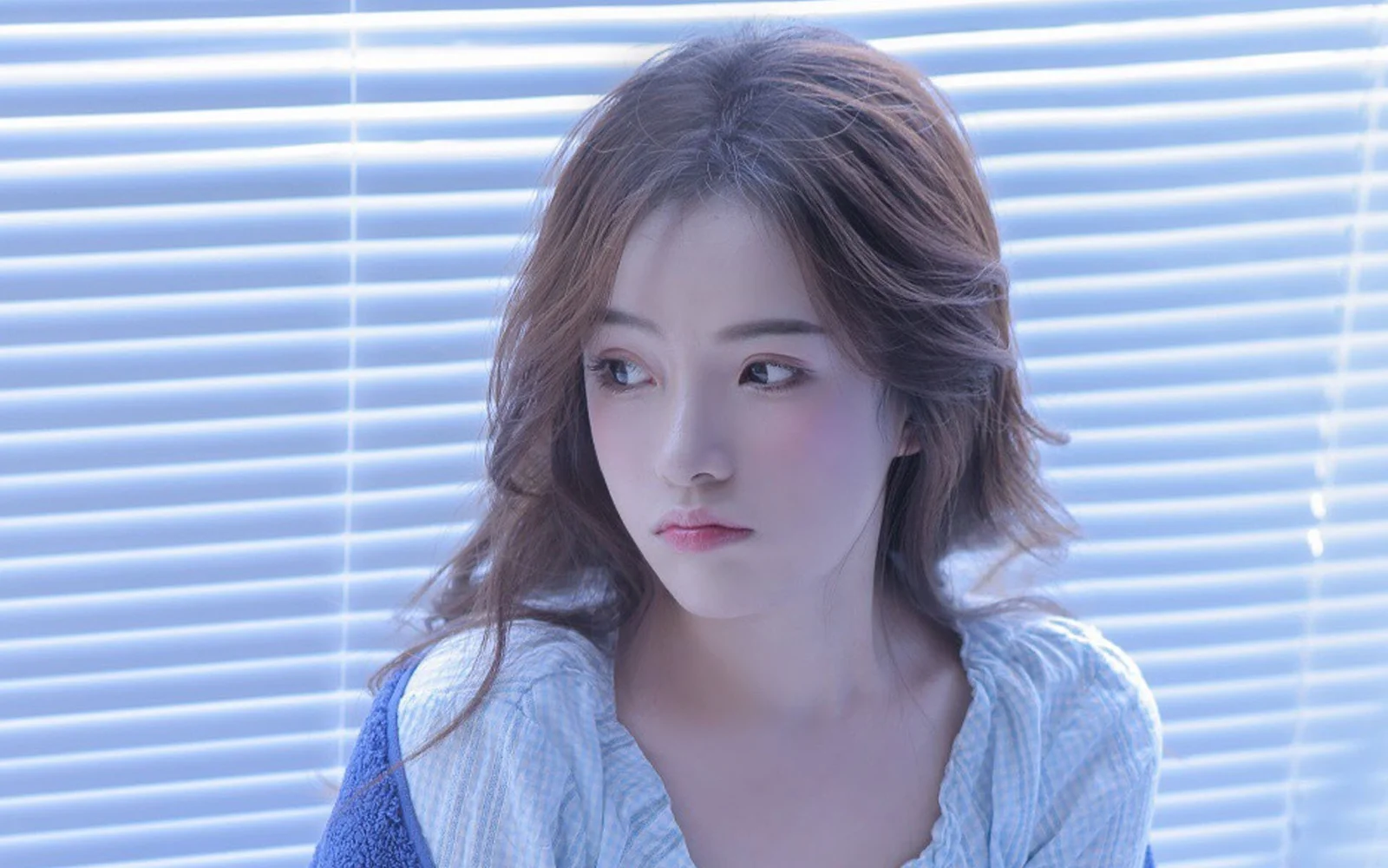 갈색 긴 머리의 여성이 수평 블라인드 앞에서 옆을 바라보며 차가운 푸른빛을 받고 있다.
