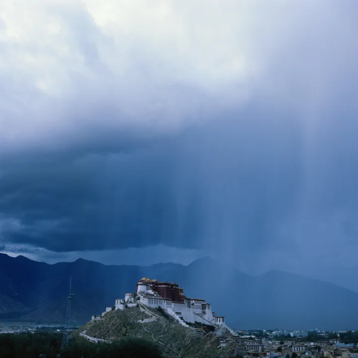 深蓝色山脉与一座山坡上的寺院建筑沐浴在阴沉云层和雨幕之中。