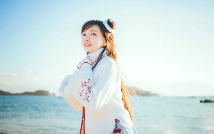 一名长发女子身穿白色上衣站在海边，阳光从侧后方照亮她的身影。