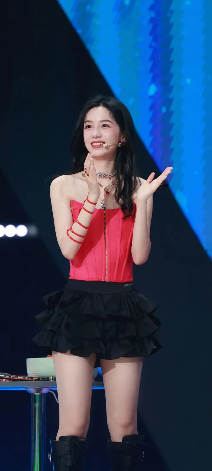 一位身穿红色上衣和黑色短裙的女性站在舞台上微笑表演。