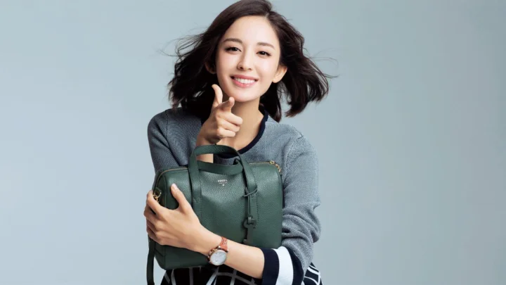 연한 회청색 배경 앞에서 성인 여성이 짙은 녹색 핸드백을 안고 미소 짓고 있습니다.