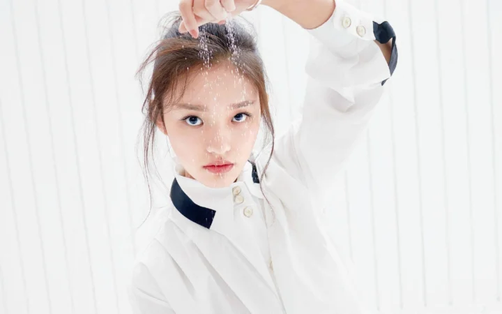 흰 셔츠를 입은 성인 여성이 밝은 흰색 배경 앞에서 한쪽 팔을 올리고 포즈를 취하고 있다.