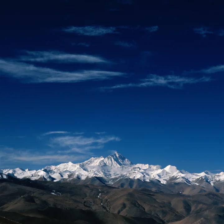 巍峨的雪山横亘在深蓝色天空下，前景是起伏的深色山丘。