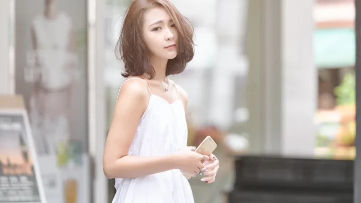 白いキャミソールドレスを着た女性が、明るい店先でスマートフォンを手に立っている。