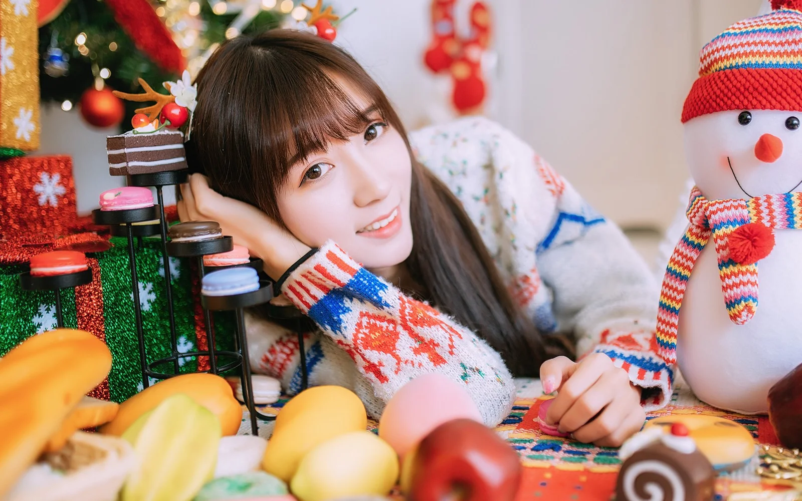 女性がクリスマスツリーや飾り、雪だるまのそばでテーブルに寄りかかりながら微笑んでいる。