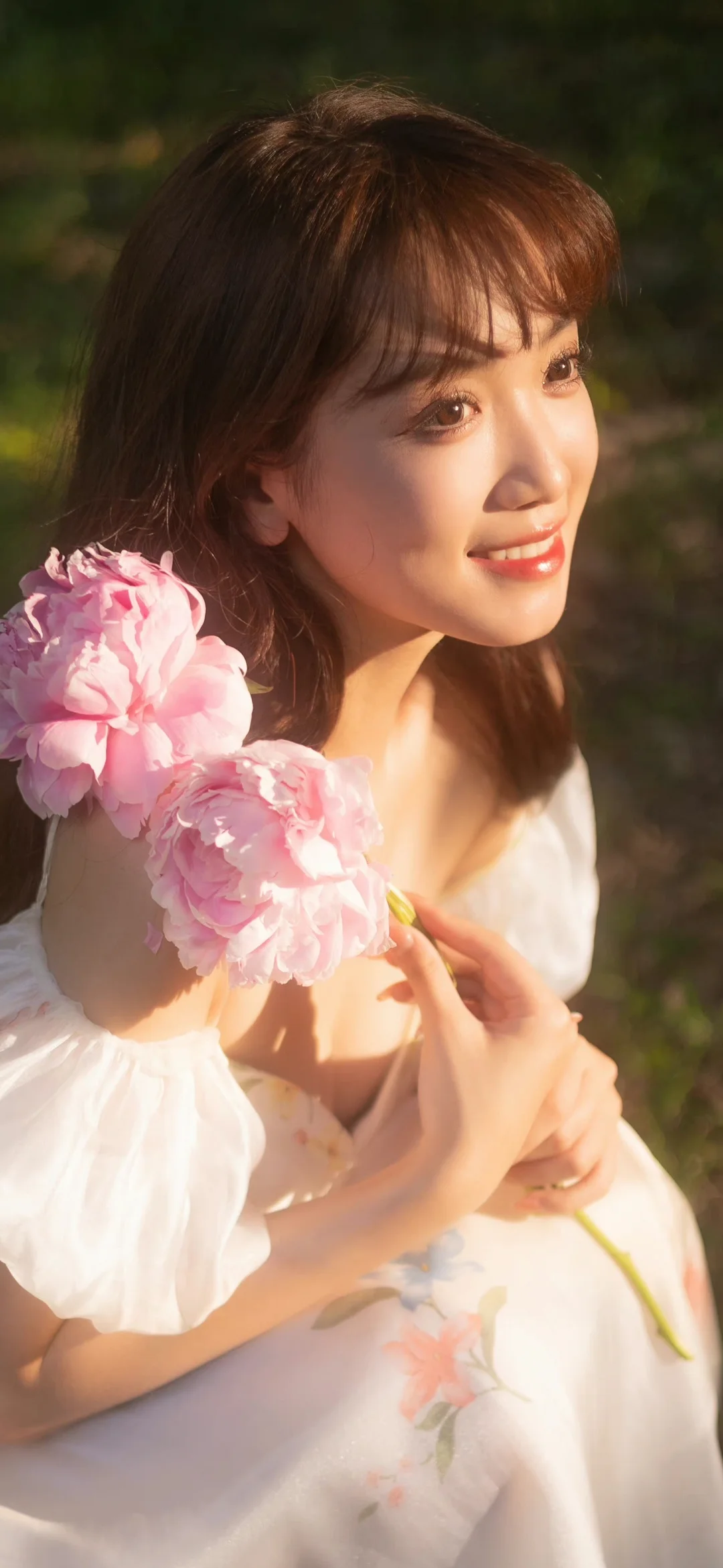 一名成年女子在户外阳光下手持粉色花朵，身穿白色连衣裙。