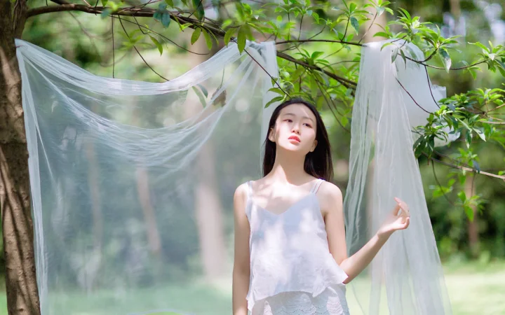 一名成年女性穿著白色吊帶上衣站在樹林中的半透明白色布幔旁。