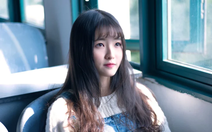 긴 머리의 여성이 실내 창가에 앉아 창문으로 햇빛이 들어오고 있다.