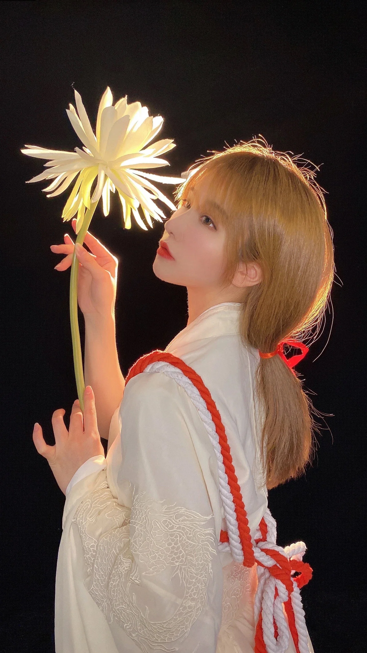 흰색 의상과 붉은 끈 장식을 착용한 여성이 검은 배경 앞에서 옆모습으로 흰 꽃을 들고 있다.