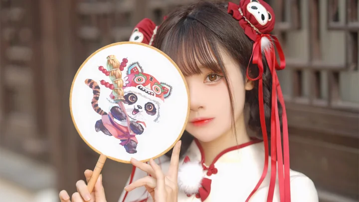 一名女性手持绣花圆扇，面部部分被扇子遮挡，红色发饰和飘带垂在两侧。