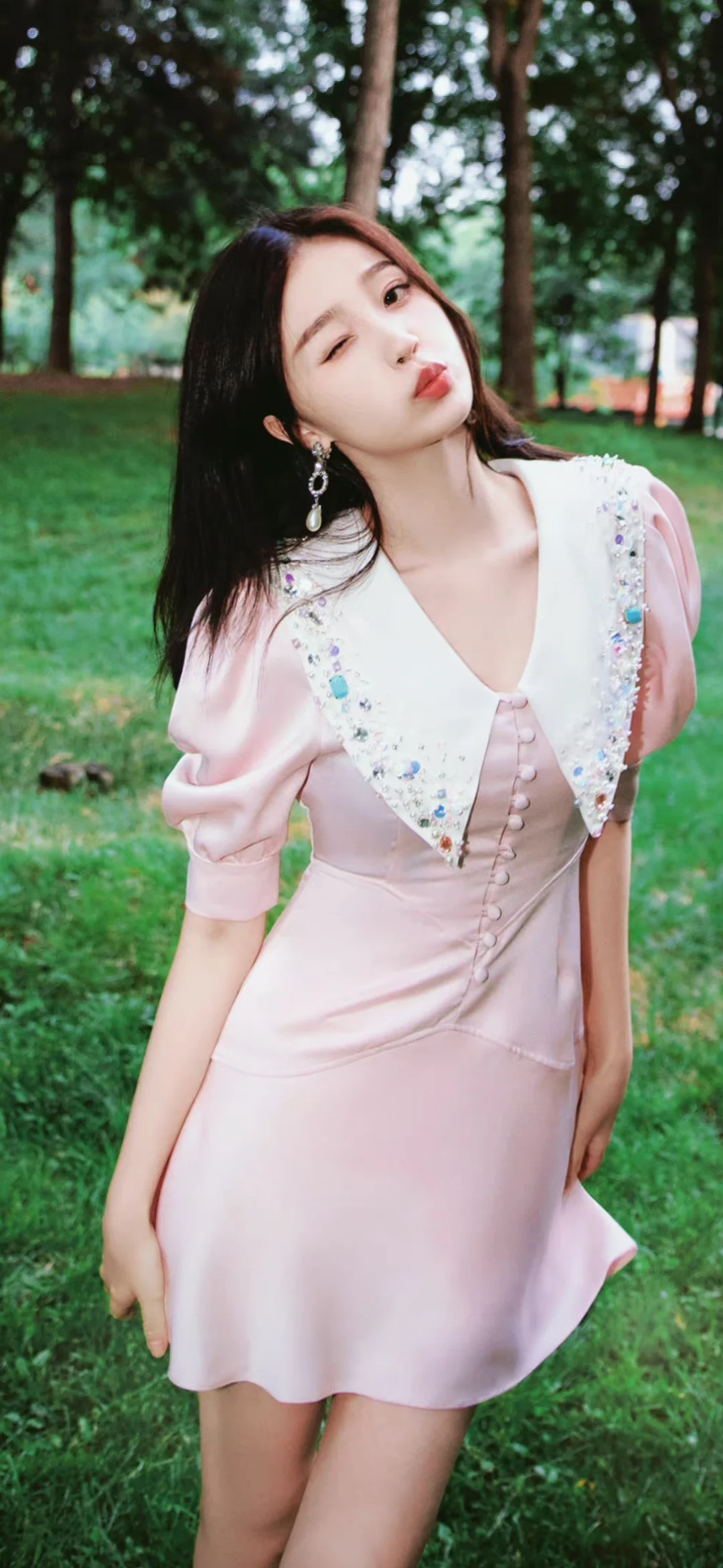 一名身穿粉色连衣裙的女性站在绿树环绕的户外草地上对镜头眨眼。