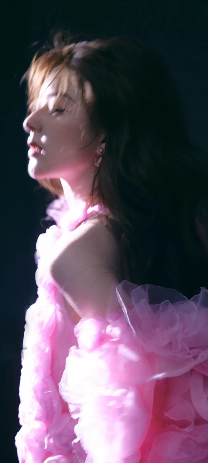 暗い背景の前で、ピンクのフリルドレスを着た成人女性が横顔を見せています。