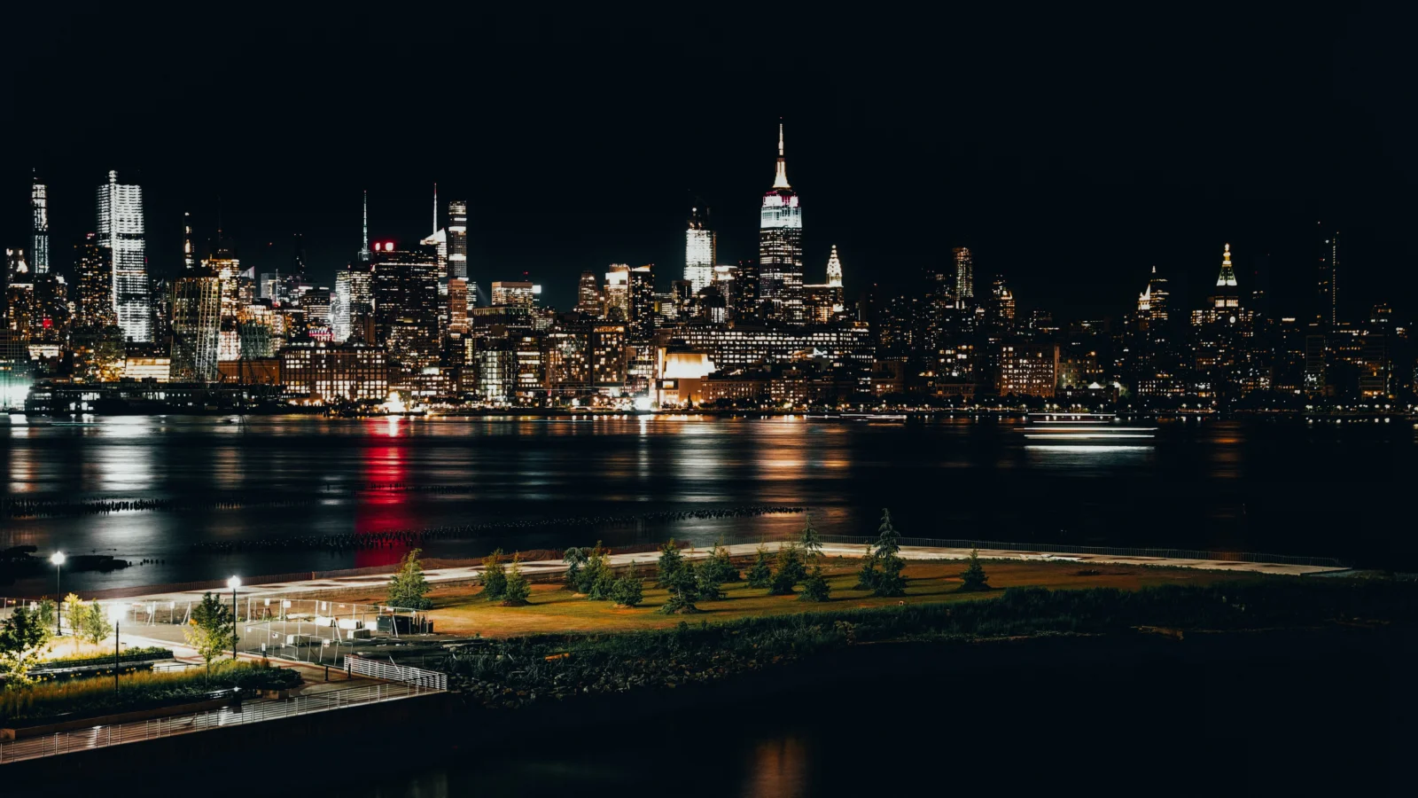 New York skyline at night across the water, with a lit park in the foreground.
