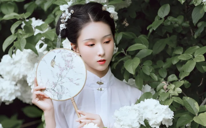 一名身穿白色传统风格服饰的女子在绿叶与白花间手持圆扇。