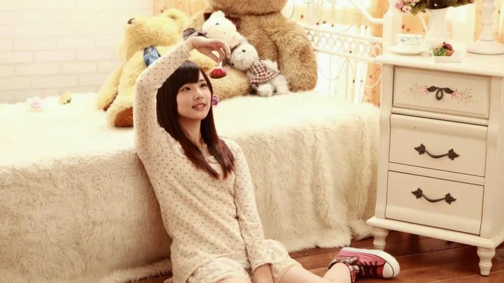 一名年轻女子坐在铺有毛绒毯的床边，身后摆放着多个泰迪熊和一组白色抽屉柜。