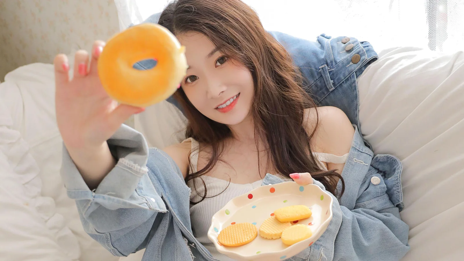 미소 짓는 성인 여성이 흰 침대 위에서 오렌지를 카메라 쪽으로 들고 있으며 앞에는 쿠키 접시가 놓여 있습니다.