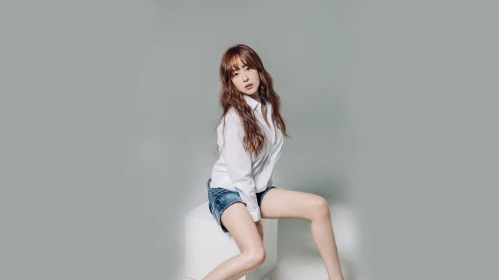 一名成年女性穿白衬衫和牛仔短裤坐在白色方台上，背景为灰色。
