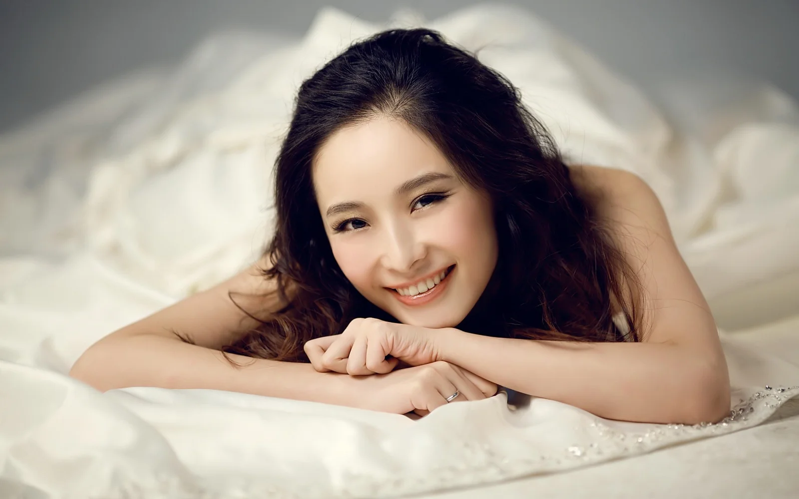 一名成年女性趴在白色床品上，微笑着将手臂交叠在身前。