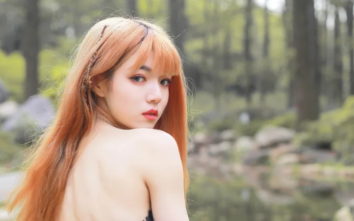 붉은 머리의 여성이 흐릿한 숲을 배경으로 어깨 너머로 카메라를 바라보고 있습니다.