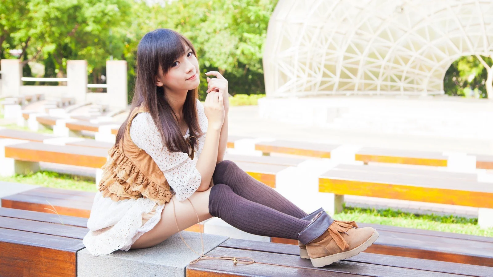一名女性坐在戶外木質長椅上，背景是綠樹和白色拱形結構。