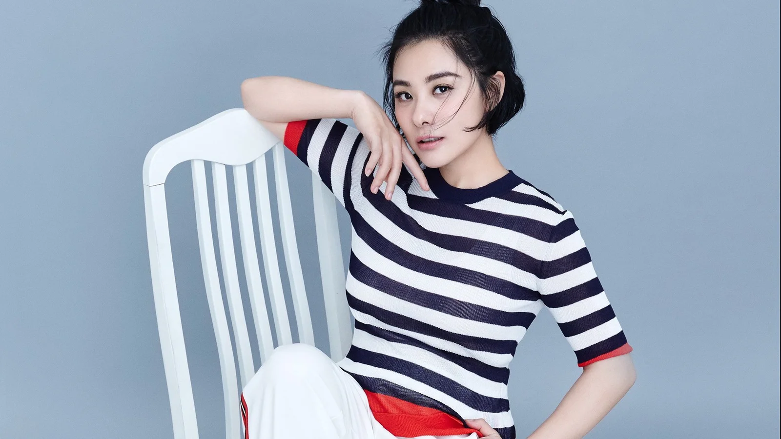 一名成年女性身穿黑白条纹上衣，倚靠在白色椅子旁的灰蓝色背景前。