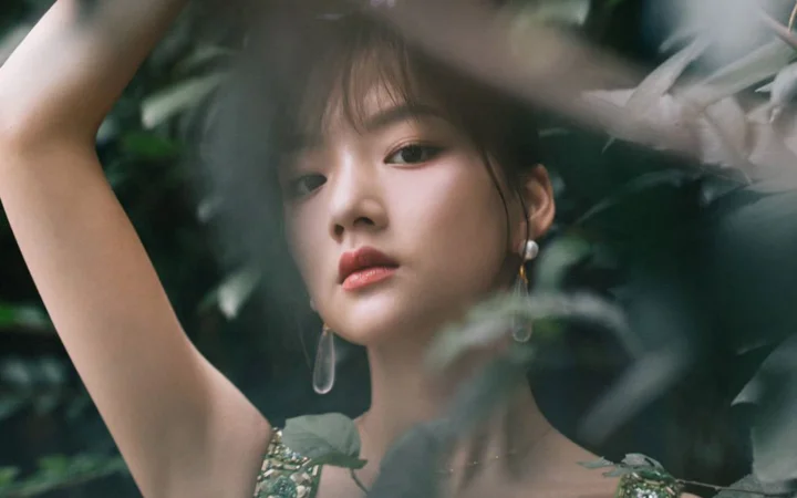 한 여성이 푸른 잎과 부드러운 빛 사이에서 팔을 들고 카메라를 바라보고 있다.