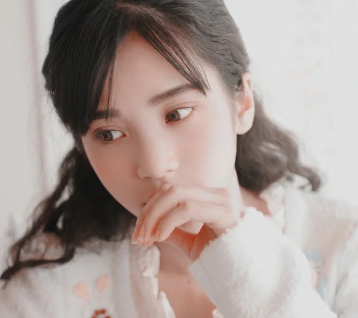 柔らかな光の中で、白いニットを着た成人女性が手を口元に添えているクローズアップポートレート。