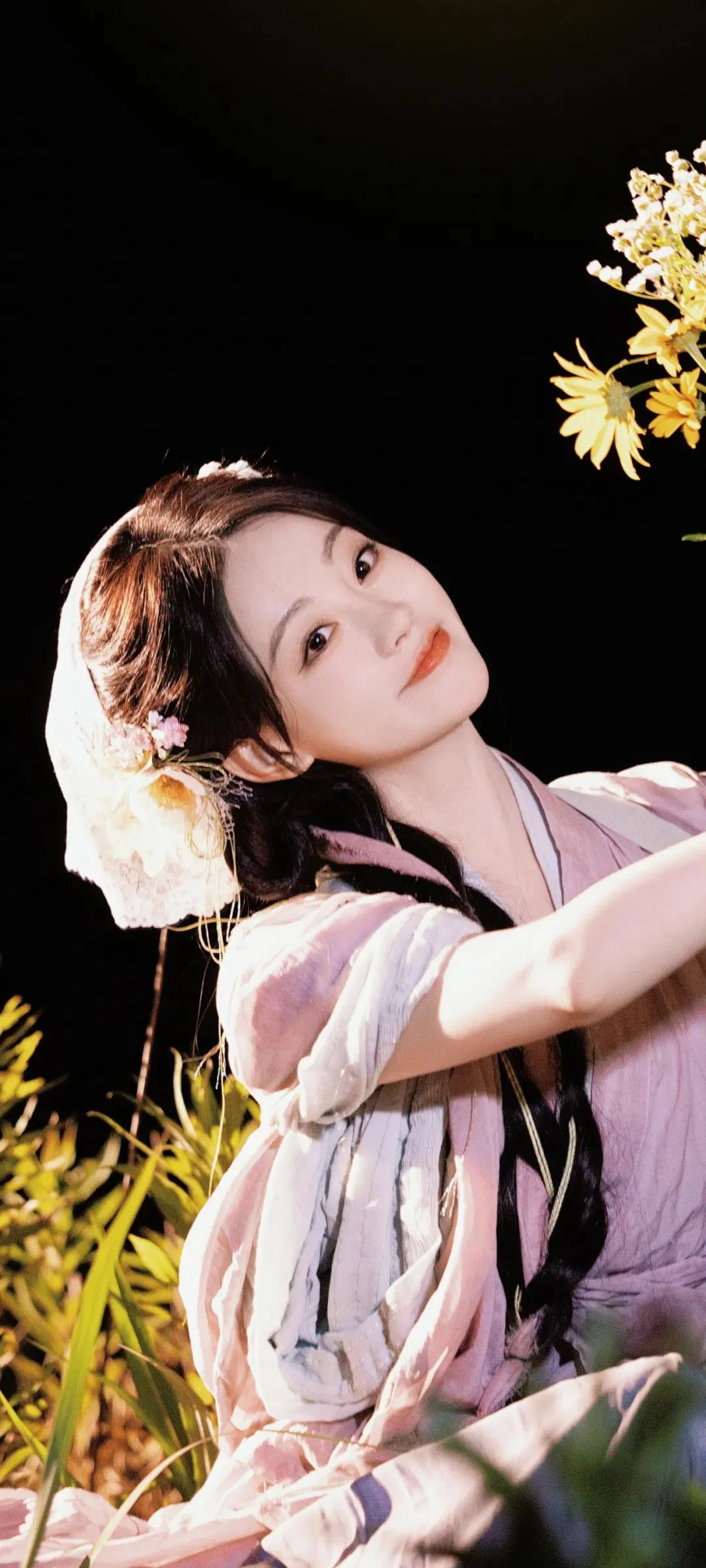 연한 전통 의상을 입은 여성이 꽃과 초목 사이에서 옆모습으로 카메라를 바라보고 있습니다.