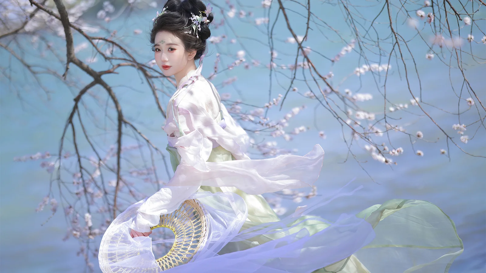 一名身穿白色传统服饰、手持团扇的女子站在蓝天下的花树旁。