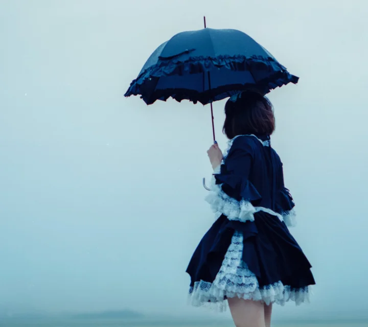 검은색 드레스를 입은 인물이 연한 파란색 배경 앞에서 등을 보인 채 짙은 파란색 우산을 들고 서 있습니다.