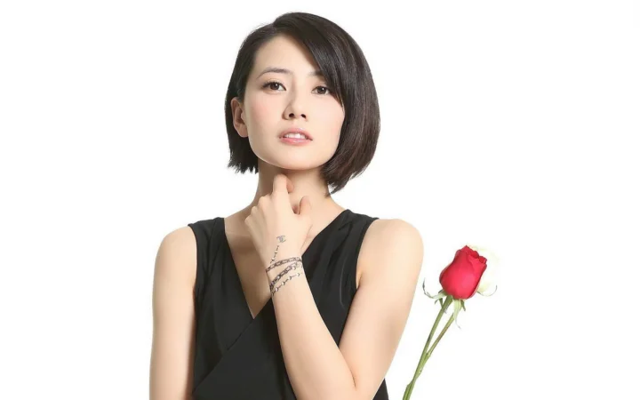 一名短发女子穿着黑色无袖上衣，手持一枝红玫瑰站在白色背景前。