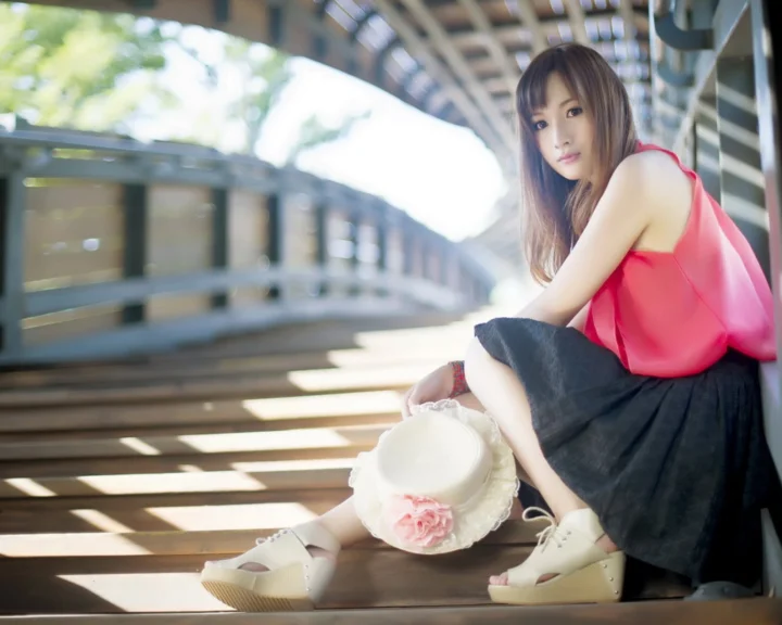一名穿粉色上衣和黑色裙子的女子坐在阳光洒落的建筑台阶上，身旁放着一顶白色花饰帽子。