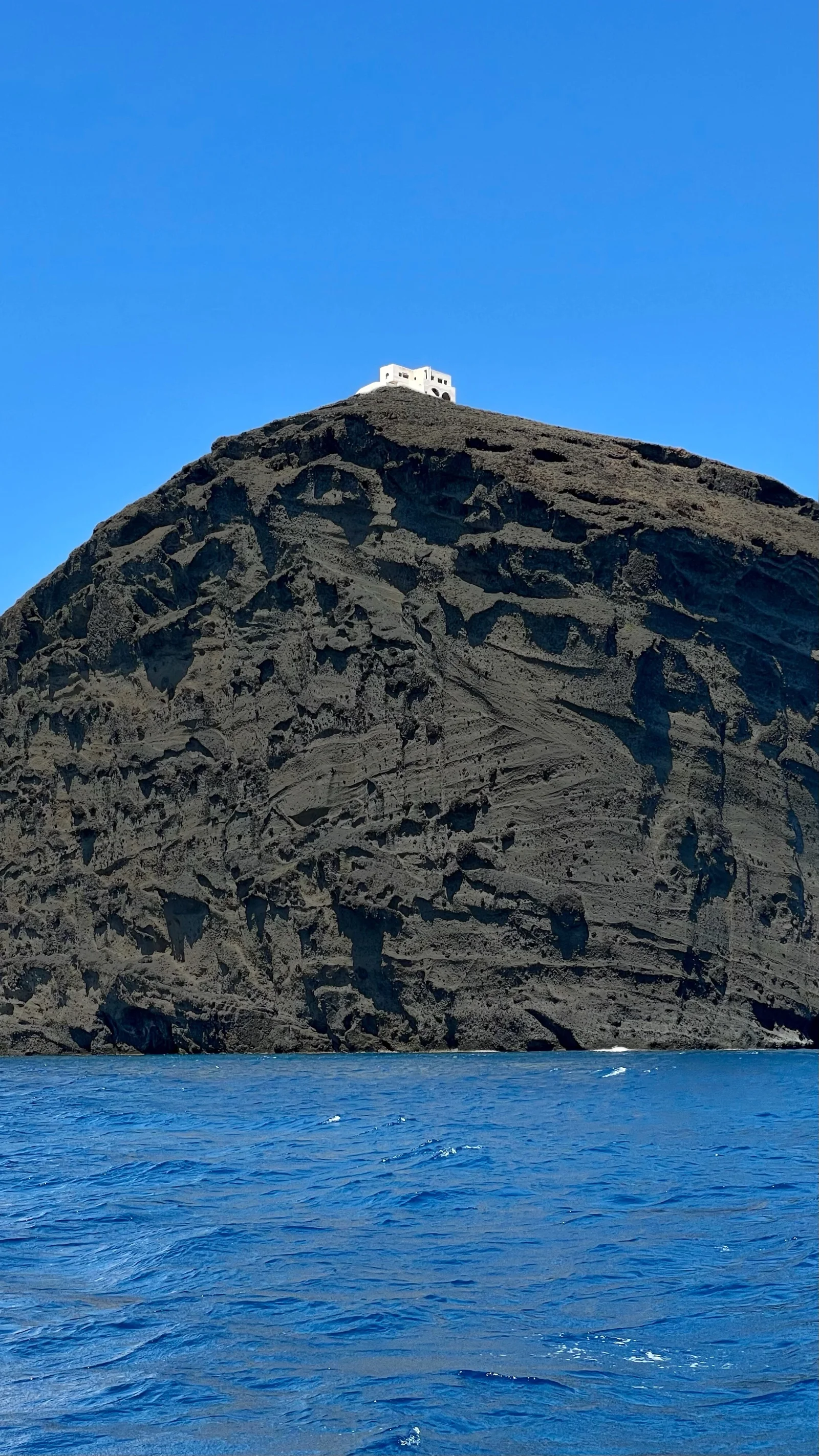 蓝色海水映衬着陡峭的深色火山岩壁，山顶可见一座白色建筑，天空晴朗无云。
