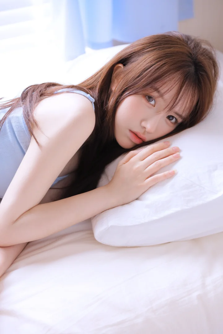 茶色の長い髪の女性が白いベッドに横たわり、枕元に腕を置いている。