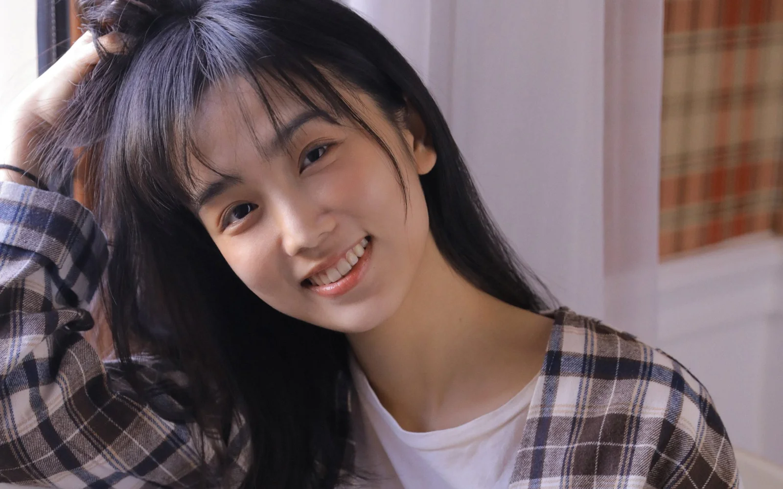 검은 긴 머리의 여성이 체크무늬 셔츠를 입고 카메라를 향해 미소 짓고 있다.