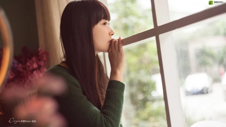 一名穿深色针织衫的女性站在窗边侧身望向窗外。