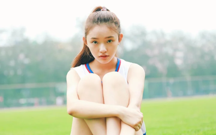 一名女子抱膝坐在草地上，身后是朦胧的树木和水面。