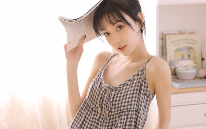 一位成年女性手持书本站在明亮的室内窗边。